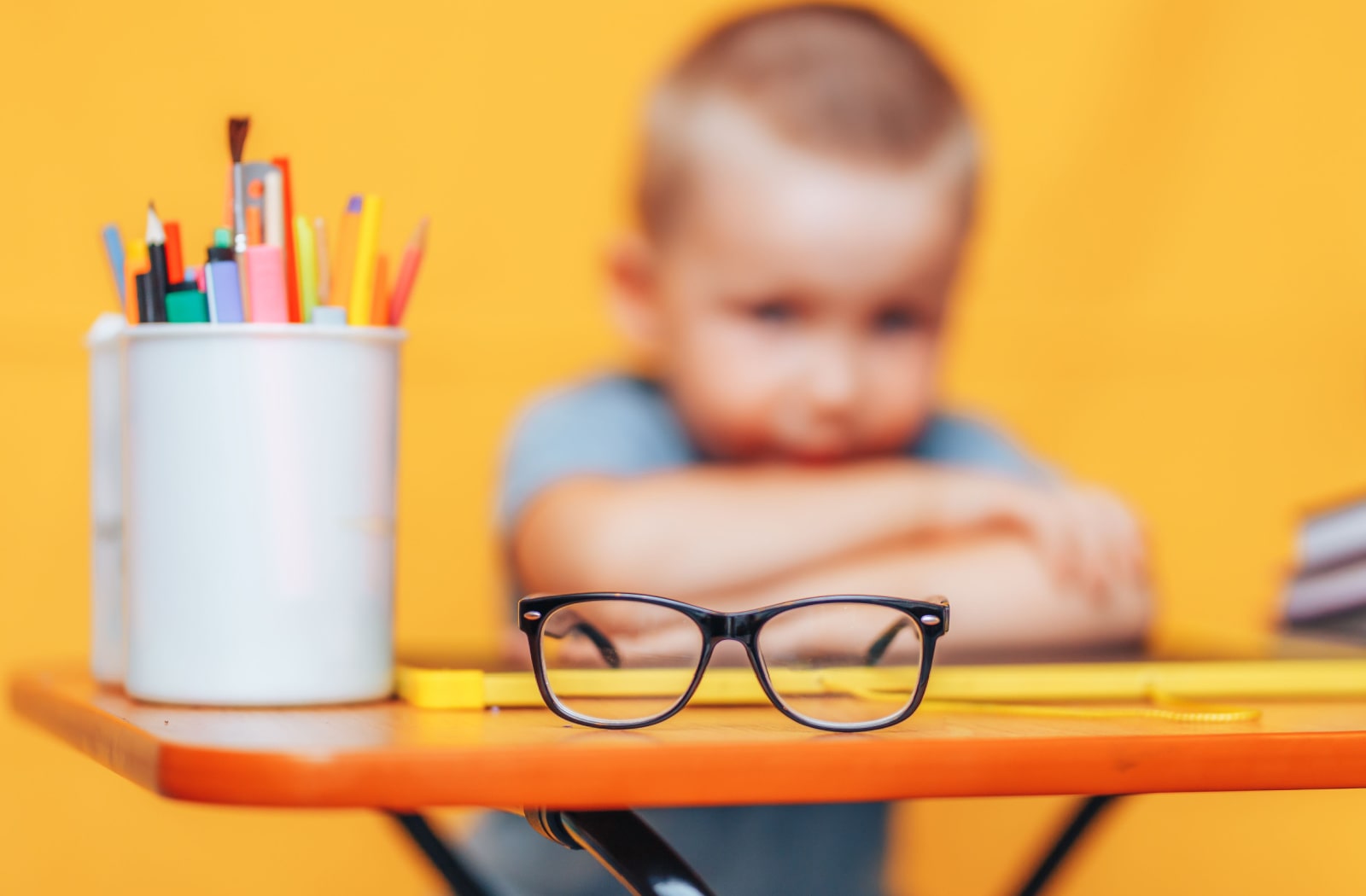 An image of a pair of corrective lenses in focus, sitting on a desk, and in the background, unfocused, is a boy sitting at the desk with his arms crossed.