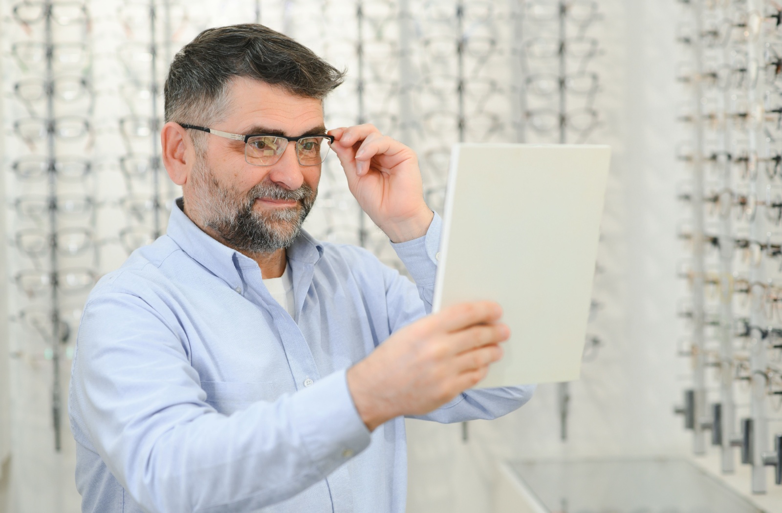 A senior holds a mirror to see themselves in a new pair of eyeglasses when getting an updated prescription at the optometrist.