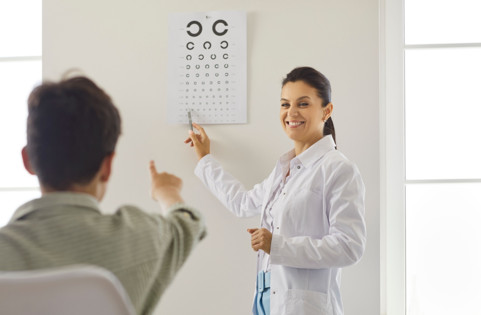 An optometrist smiles while pointing to a vision chart as a child identifies the letters.