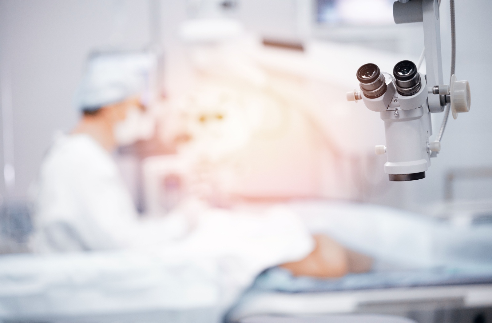 A surgical microscope is suspended in the foreground while a blurred medical professional performs LASIK in the background.